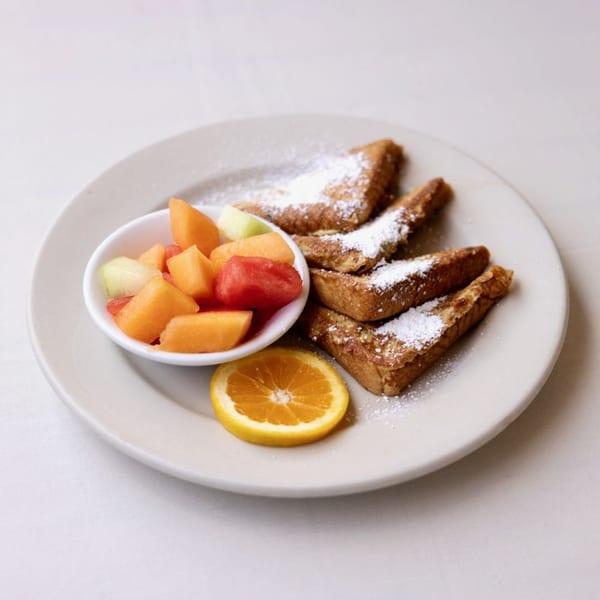 French toast dusted with powdered sugar, a side of fresh fruit, and an orange slice on a white plate.