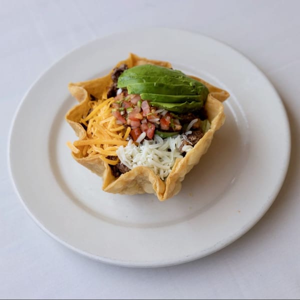 A taco salad in a crispy tortilla bowl, topped with avocado, pico de gallo, and shredded cheese.